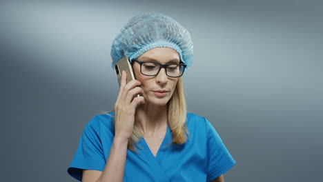 Young-Caucasan-busy-blonde-woman-doctor-in-blue-costume,-hat-and-glasses-talking-on-the-phone-emotionally.-Close-up.